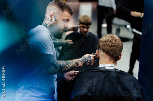 Male haircut with electric razor. Tattooed Barber makes haircut for client at the barber shop by using hairclipper. Man hairdressing with electric shaver.