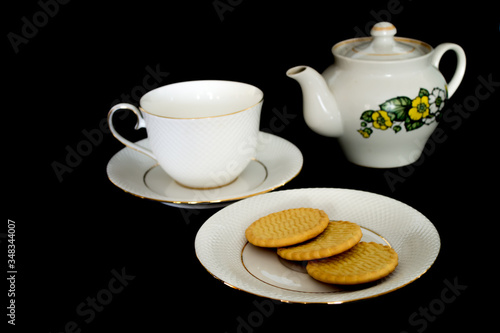 The tea set, consisting of a tea cup with a saucer, a kettle and a plate of biscuits, is located on a black background.