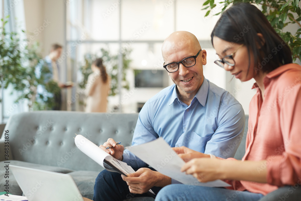 Portrait of bald mature businessman smiling at Asian young woman while discussing project in modern office, copy space Portrait of bald mature businessman smiling at Asian young woman while discussing project in modern office, copy space