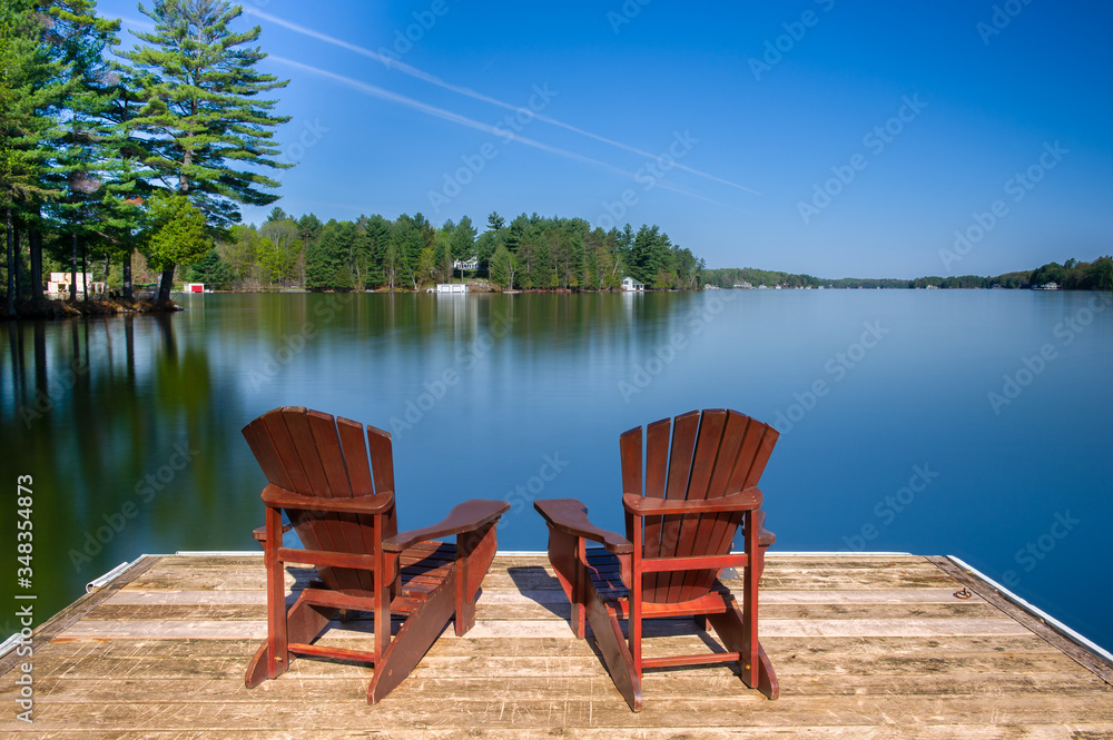 Two Muskoka chairs sitting on a wood dock facing a lake. Across the calm water is a white cottage nestled among green trees. There are no clouds in the sky.
