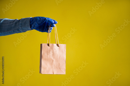 Closeup of female hands in gloves and a denim shirt. A delivery man is holding a small paper bag by the pens on a yellow background. Craft packaging for takeaway snack. Antimicrobial protection.