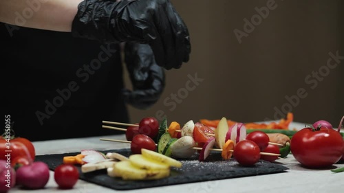 Woman in black gloves sprinkles salt kebab of vegetables on a wooden table. Preparing healthy food