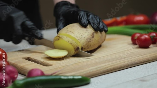 Female hands in black gloves with a sharp knife cutting the potatoes on the wooden board in the kitchen.