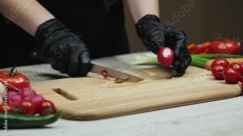 Hands in black gloves slices red radish on wooden board in the kitchen