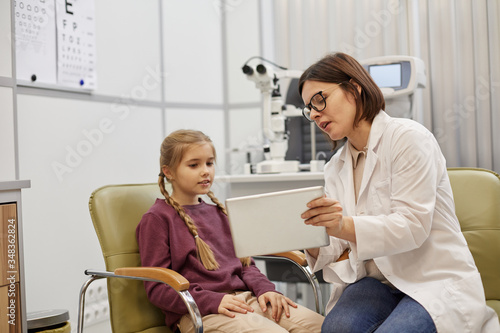 Portrait of young female ophthalmologist using digital tablet while talking to little girl during consultation in modern clinic, copy space photo