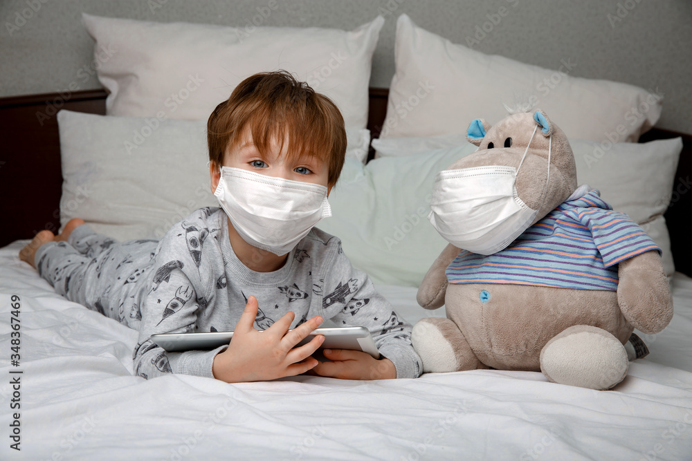 Sick boy in a medical protective mask uses a tablet computer lying on a bed at home . Concept of quarantine and coronavirus epidemic.