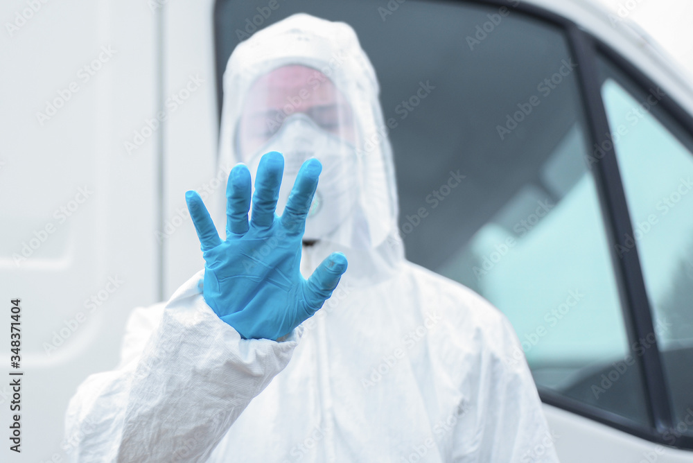 Tired Doctor ambulance in a protective antivirus suit, a face and eye mask with gloves, stands next to the ambulance, With downcast eyes and a raised hand, Sweeping palm indicating stop signal