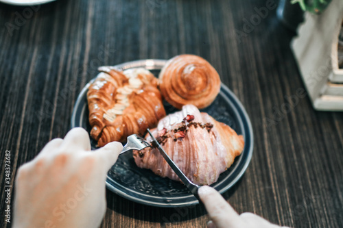 Hands prepare for eating fresh sweet bake croissant almond by use a fork and knife for cutting on a black plate for breakfast over on dark wooden table background morning time.copy space for your text