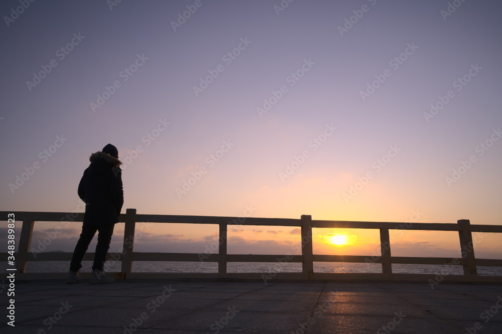 Silhouette of sad man standing before fence by the sea, low angle view, sunset background, copy space
