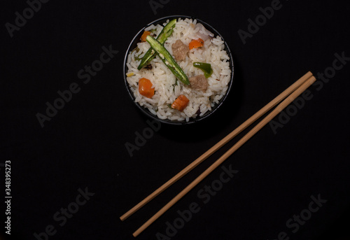 Top down image of a bowl of rice with vegetables and soya chunks along with chopsticks in a black copy space background. Food photography.