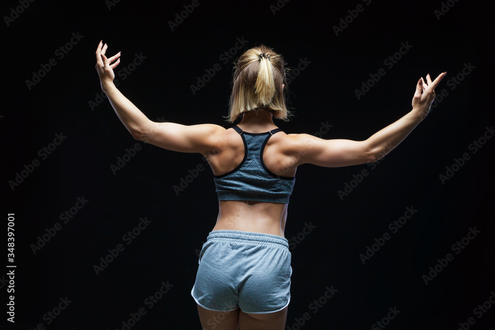 Back view of strong sporty girl in top and jean shorts showing muscles on black background