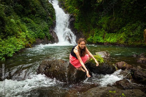 Young Caucasian woman sitting on the rock and playing with water in the river near the waterfall. Travel lifestyle. Dedari waterfall in Sambangan  Bali.