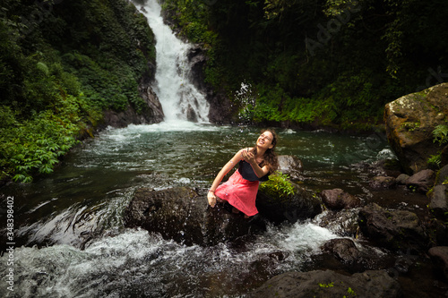 Happy Caucasian woman sitting on the rock and playing with water in the river near the waterfall. Travel lifestyle. Dedari waterfall in Sambangan  Bali.