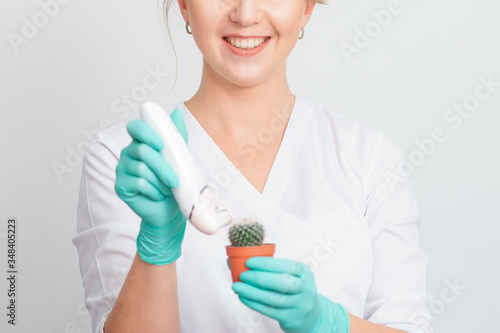 Cosmetologist is shaving green cactus in a brown pot with electric epilator wearing gloves smiling on white background. Concept of depilation  epilation and removal hair.