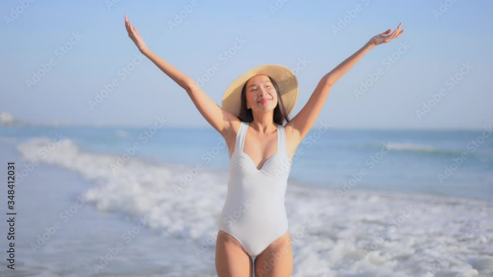 Static shot of a beautiful sexy lady wearing a white swimsuit and hat happy standing on the beach with her hands up in excitement Static shot of a beautiful sexy lady wearing a white swimsuit and hat happy standing on the beach with her hands up in excitement