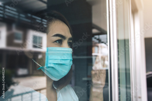 Asian woman looking through the window and wearing medical mask protection from the illness with stay isolated quarantine COVID-19 pandemic virus mask against coronavirus disease at home.