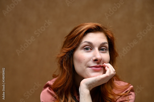 Portrait of actress caucasian woman with red hair showing joyful emotions. Photo taken in the studio on a beige background. The model is wearing a red shirt.