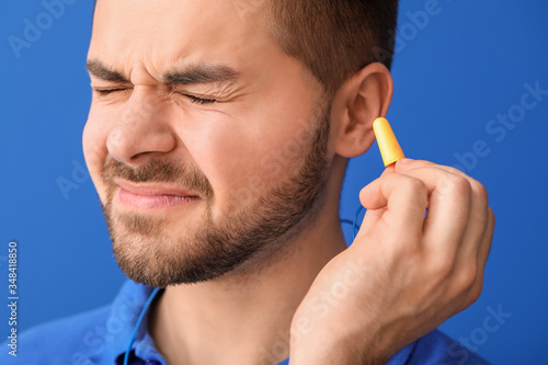 Young man with ear plugs suffering from loud noise on color background