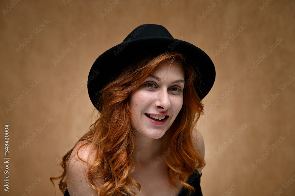 Studio large photo portrait of a caucasian woman with long red hair on a beige background. Model posing with a smile in a hat.