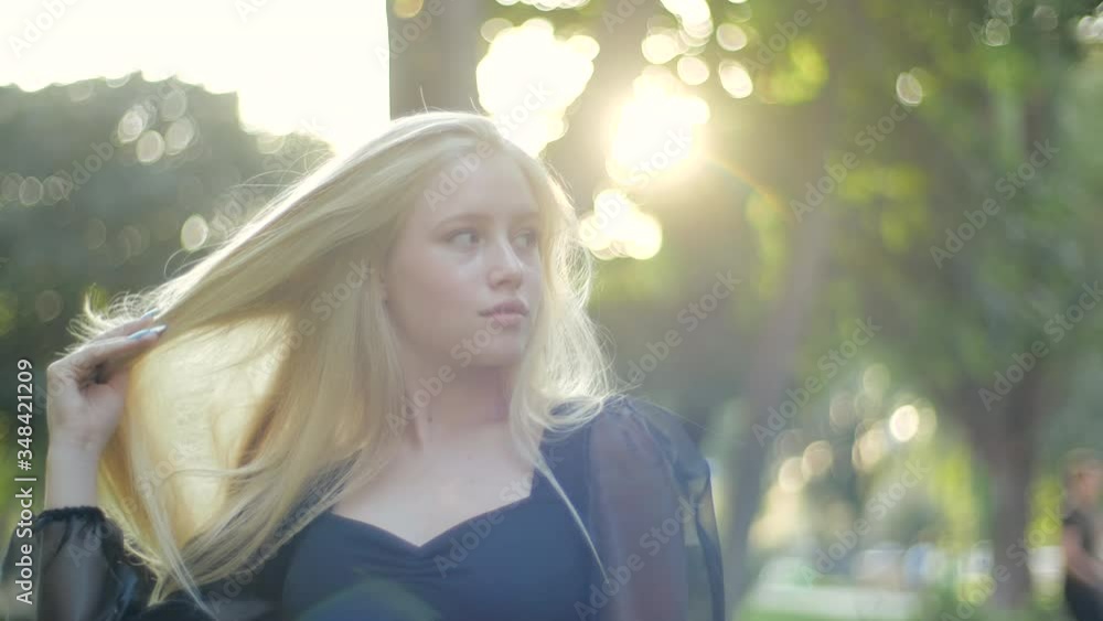 A young attractive Caucasian girl turns, looks at the camera and straightens her long blond healthy hair. Chic fashion teenager model smiling confidently and posing outdoors in the park. A young attractive Caucasian girl turns, looks at the camera and straightens her long blond healthy hair. Chic fashion teenager model smiling confidently and posing outdoors in the park.