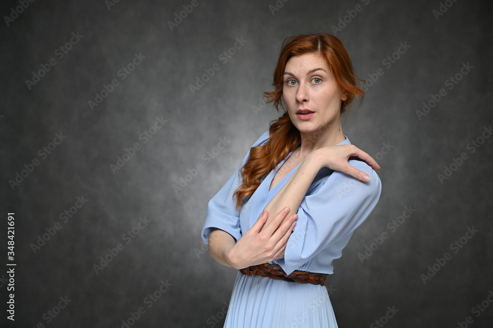 Portrait of a young caucasian woman with long red hair in a blue dress. The model poses with different emotions on a gray background in the studio. Portrait of a young caucasian woman with long red hair in a blue dress. The model poses with different emotions on a gray background in the studio.
