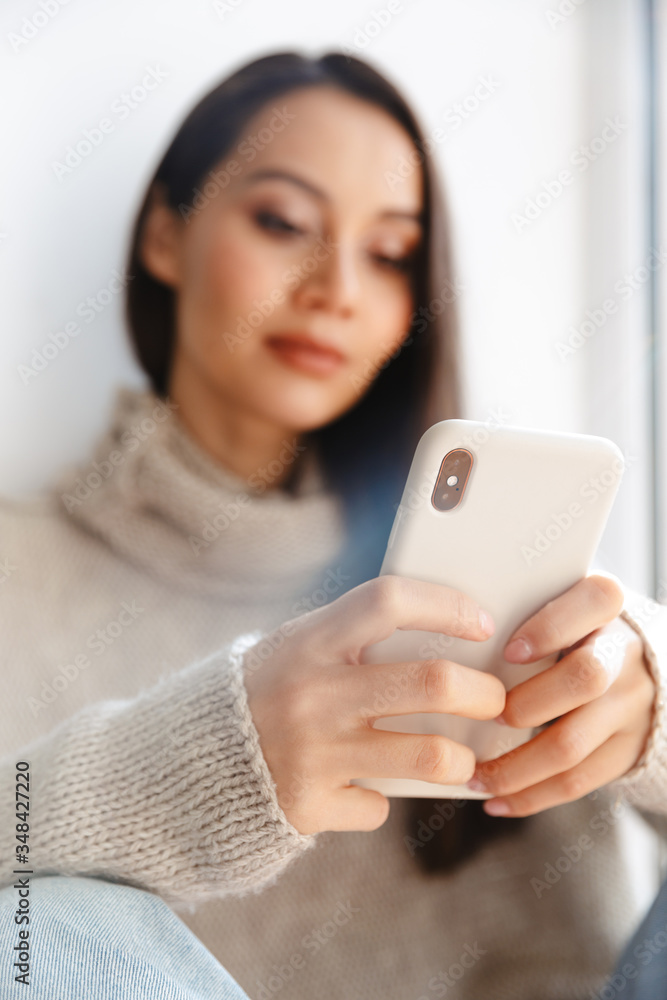 Image of asian woman typing on mobile phone while sitting on window sill Image of asian woman typing on mobile phone while sitting on window sill