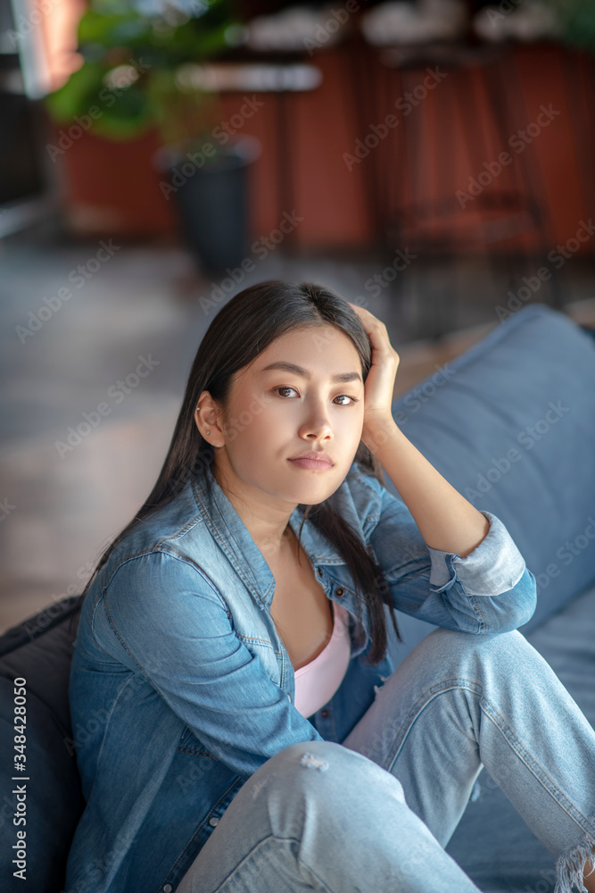 Fototapeta premium Upset dark-haired female sitting with folded knees on sofa, thinking, resting her head on one hand