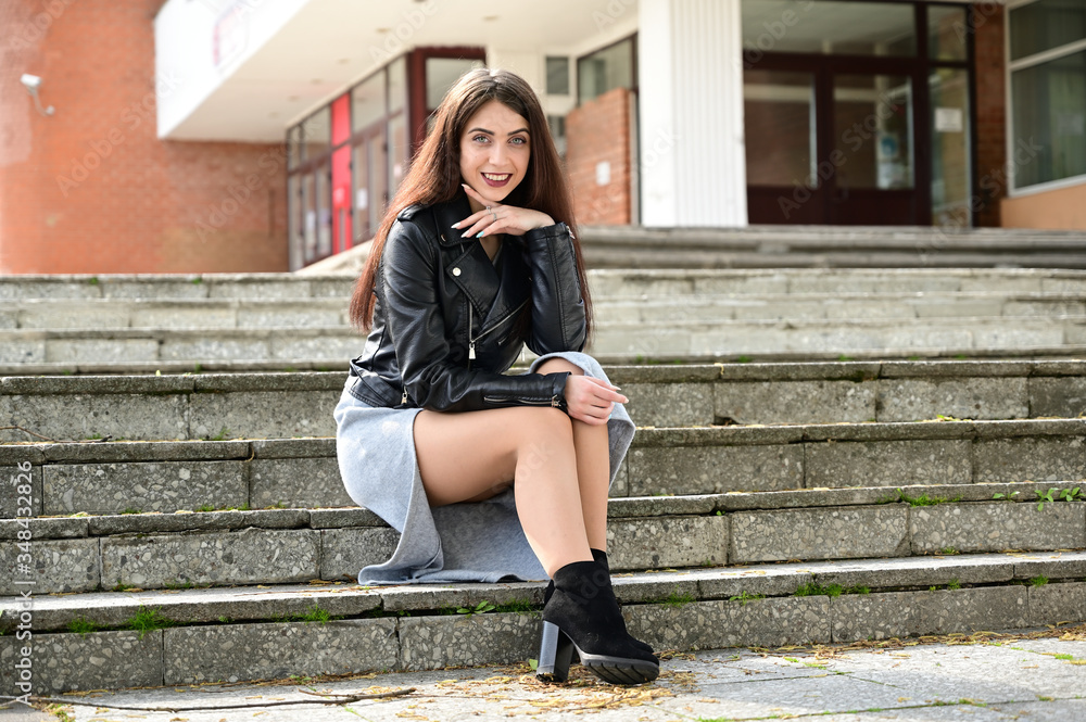 Full-length photo of a pretty young Caucasian brunette girl in a black jacket sitting on the steps of the building in different poses. Model posing in the city in the spring outdoors with a smile. Full-length photo of a pretty young Caucasian brunette girl in a black jacket sitting on the steps of the building in different poses. Model posing in the city in the spring outdoors with a smile.