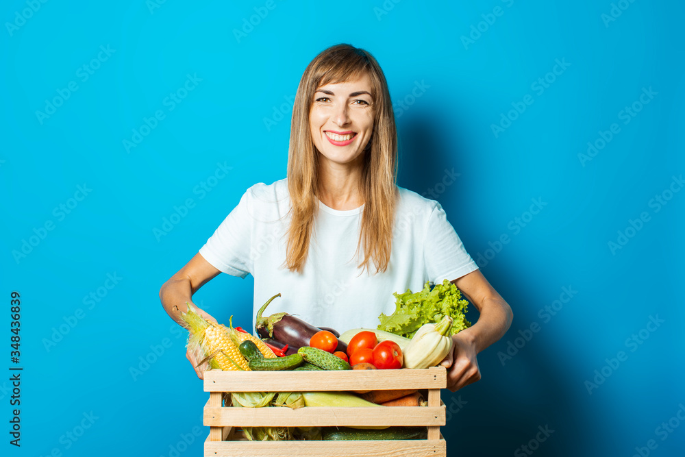 Young woman holds a box with fresh vegetables on a blue background. Good harvest concept, natural product