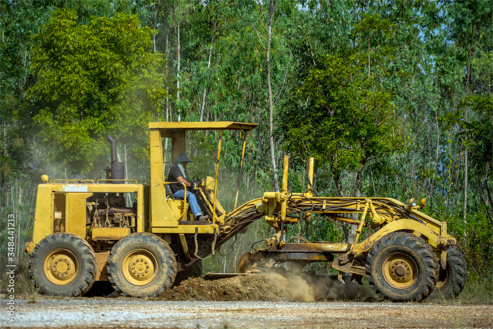 Motor grader clearing and leveling construction site surface with forest in the background. Grader industrial machine on road construction work. Motor grader clearing and leveling construction site surface with forest in the background. Grader industrial machine on road construction work.