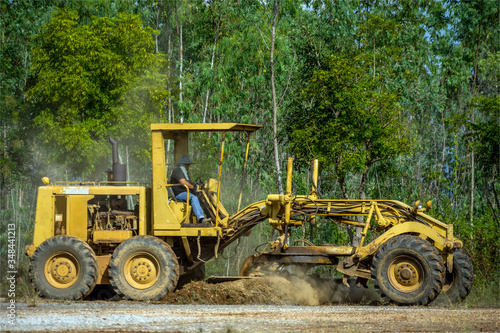 Motor grader clearing and leveling construction site surface with forest in the background. Grader industrial machine on road construction work.