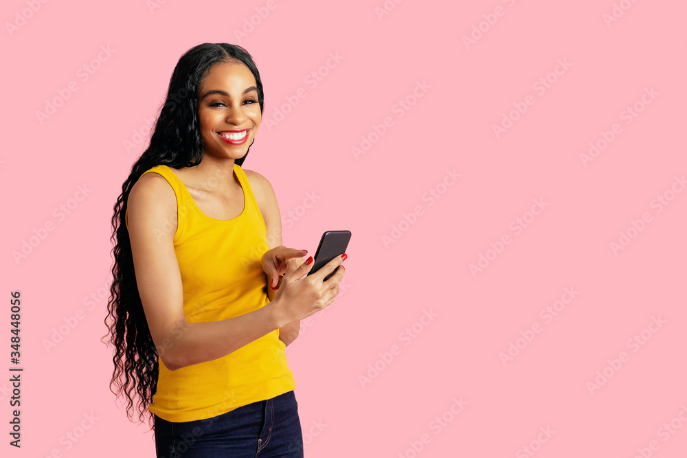 Portrait of a smiling young woman with long black curly hair pressing and using smart phone, against pink studio copy space Portrait of a smiling young woman with long black curly hair pressing and using smart phone, against pink studio copy space