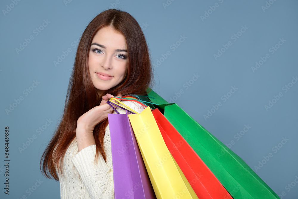 Beautiful happy smiling satisfied young woman with long ginger hair holding colored shopping bags over blue background. Sale, Black Friday, purchases, consumption concept Beautiful happy smiling satisfied young woman with long ginger hair holding colored shopping bags over blue background. Sale, Black Friday, purchases, consumption concept