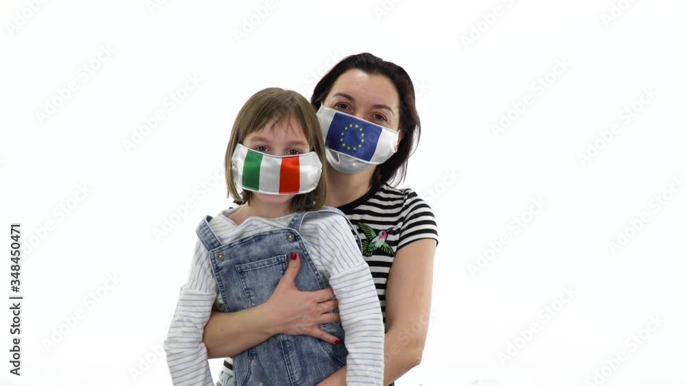 Mom and daughter posing on a white background. Quarantine, virus and pandemic concept. On a woman and a child a protective medical mask with the flags of Italy and the European Union, Europe. Mom and daughter posing on a white background. Quarantine, virus and pandemic concept. On a woman and a child a protective medical mask with the flags of Italy and the European Union, Europe.