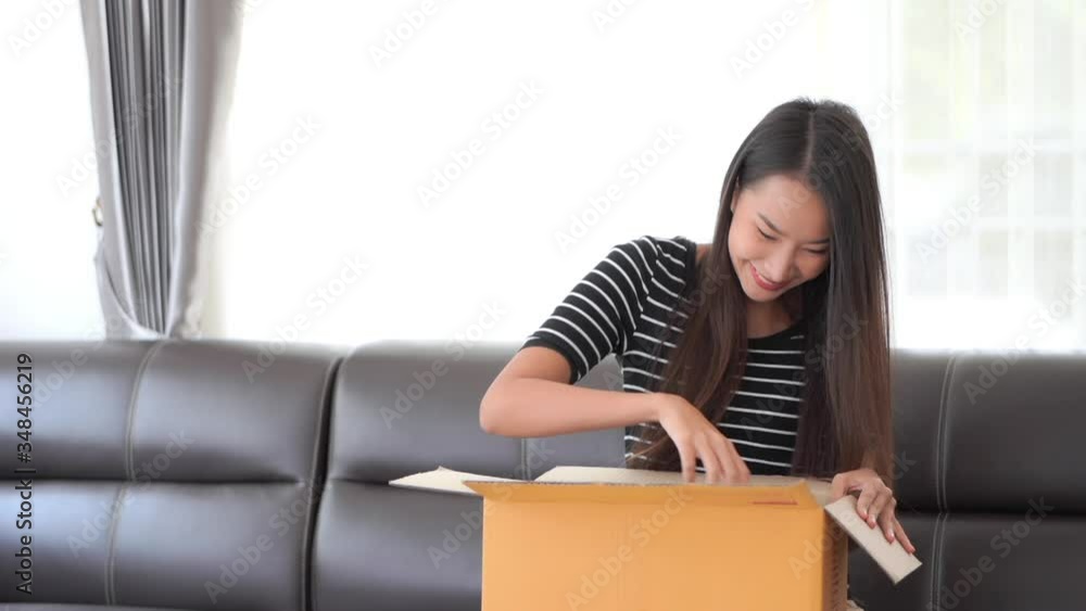 Happy Asian Female Opens Present Box While Sitting on Sofa in Family Home Happy Asian Female Opens Present Box While Sitting on Sofa in Family Home