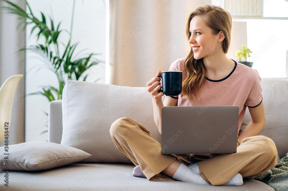 Young attractive girl with a cup of coffee sitting on a sofa holding a laptop, looking to the side and smiling. Online chatting Young attractive girl with a cup of coffee sitting on a sofa holding a laptop, looking to the side and smiling. Online chatting