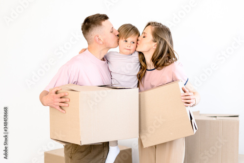 Happy family life. Young parents and their little son are standing holding cardboard boxes. They moved to their new own home and kisses their beloved son