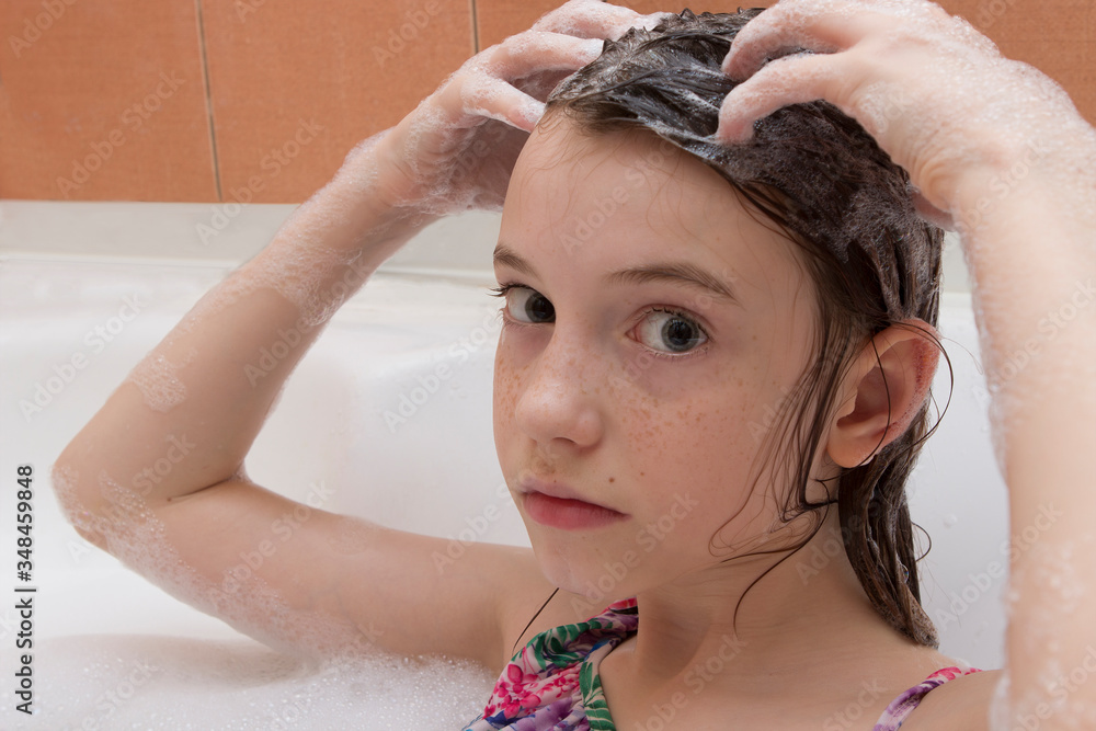 Fototapeta premium A girl with freckles in a bathing suit washes her head in the bath. Baby in bathtub with bath foam.