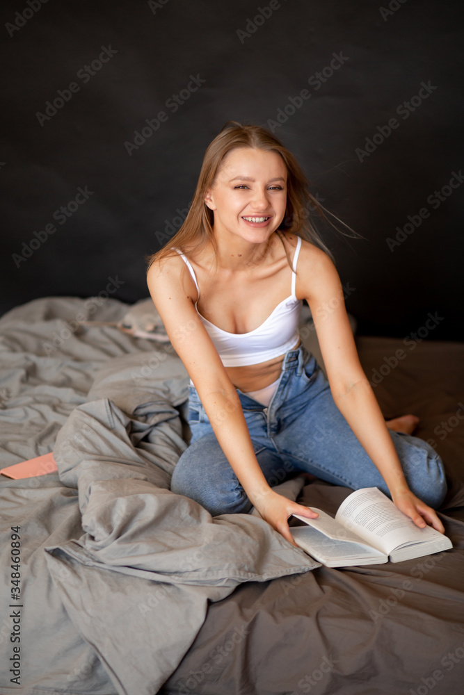 Beautiful girl in jeans and a top sits on a bed with a book, looks at the camera and smiles Beautiful girl in jeans and a top sits on a bed with a book, looks at the camera and smiles