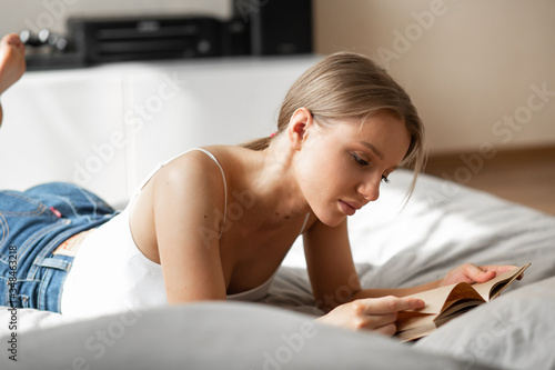 Beautiful girl in jeans and a top lies on a bed at home with a book and reads