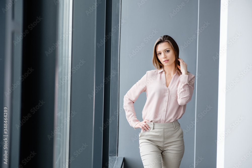 Confident young positive pretty smiling caucasian woman in business suit rejoices standing on gray background in modern office. Concept of successful career and good job Confident young positive pretty smiling caucasian woman in business suit rejoices standing on gray background in modern office. Concept of successful career and good job