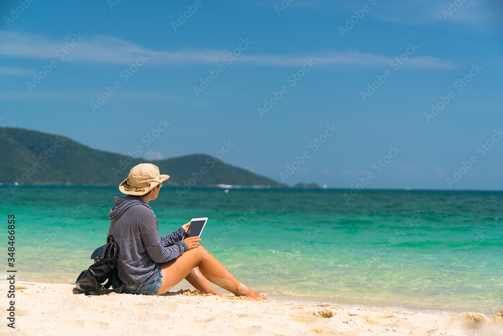 hiker girl in a hat with a backpack with glasses sitting on the sand with a tablet in hands.