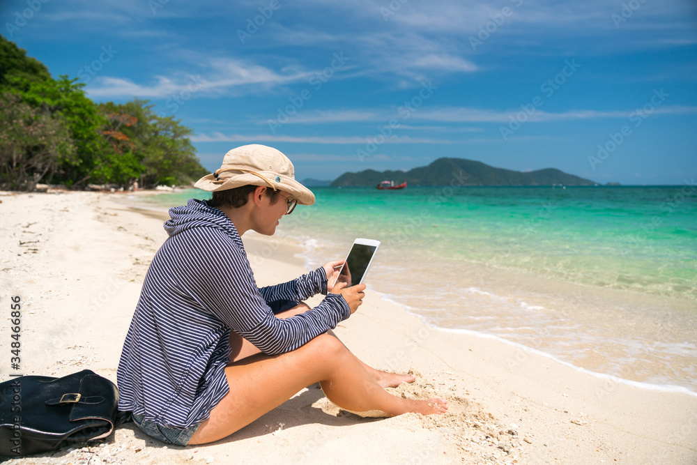 Beautiful Hiking girl with a backpack sittit on a sandy tropical beach and uses a tablet and internet for work.