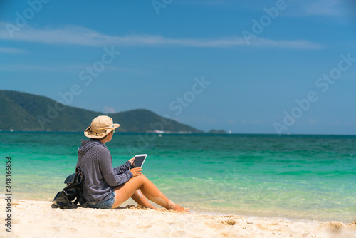 hiker girl in a hat with a backpack with glasses sitting on the sand with a tablet in hands.