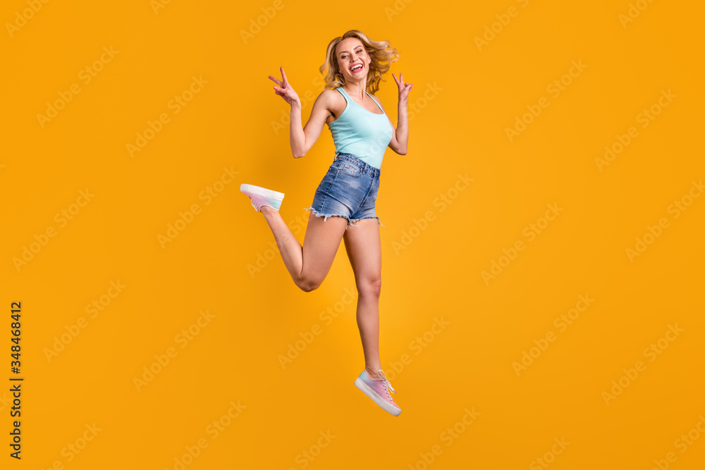 Full length profile photo of attractive pretty lady good mood jump high excited street summer look show v-sign symbol wear teal tank-top denim shorts shoes isolated yellow color background Full length profile photo of attractive pretty lady good mood jump high excited street summer look show v-sign symbol wear teal tank-top denim shorts shoes isolated yellow color background
