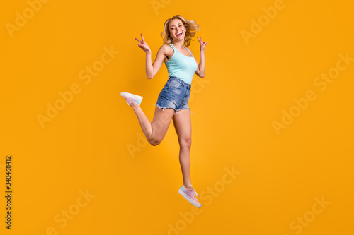 Full length profile photo of attractive pretty lady good mood jump high excited street summer look show v-sign symbol wear teal tank-top denim shorts shoes isolated yellow color background
