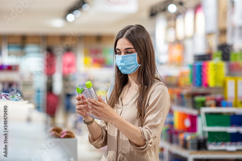 Woman wearing face mask push shopping cart in supermarket department store. Girl choosing, looking grocery things to buy at shelf during coronavirus crisis or covid19 outbreak.