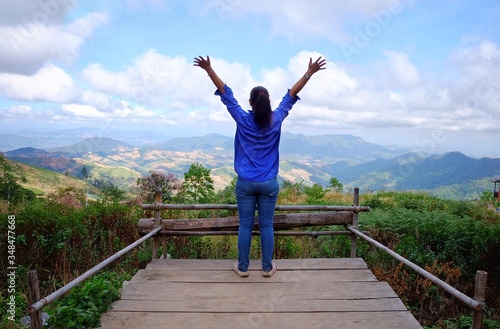 The back view of a woman with blue shirt and jeans looking over a lush green mountains and valley at the edge of a wooden ledge with her arms up  feeling happy and relax after a long journey.