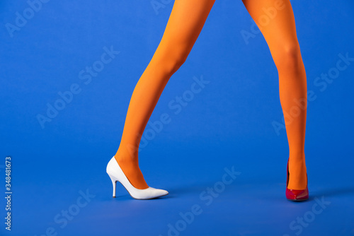 cropped view of trendy model in bright orange tights, white and red heels posing on blue