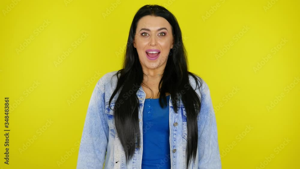 Woman with black hair in a denim jacket looks at the camera and is surprised with her mouth open on a yellow background with copy space. Place for text or product. 4K footage Woman with black hair in a denim jacket looks at the camera and is surprised with her mouth open on a yellow background with copy space. Place for text or product. 4K footage